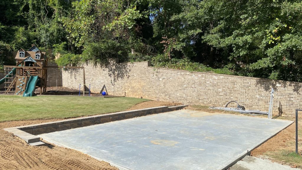 A backyard with a partially constructed concrete pad, surrounded by trees and a brick wall. A wooden playset with slides stands in the grass.